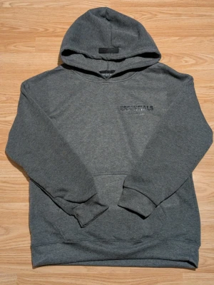 Grå Essentials Fear of God hoodie - Mörkgrå hoodie från Essentials Fear of God med huva och tryck på både bröstet och ryggen. Klassisk passform med ribbade muddar vid ärmar och nederkant. Tillverkad i mjukt bomullsmaterial för maximal komfort. Perfekt för en avslappnad streetwear-look.
