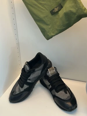 Svarta sneakers från Äkta - Snygga svarta sneakers med grå och svart camomönstrad mesh och svarta detaljer i mocka och läder. Klassisk låg modell med svart sula och svarta skosnören. Perfekta för dig som vill ha en stilren men ändå unik look.