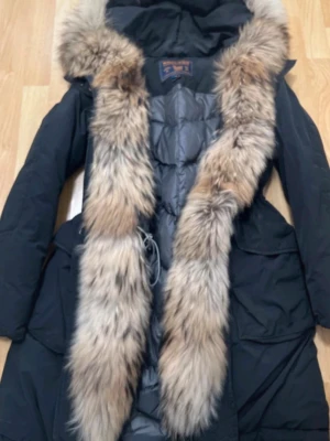Woolrich Svart parkas med äkta pälsdetalj - Snygg svart parkas från Woolrich med stor luva och lyxig äkta päls runt huvan. Jackan har quiltat innerfoder och dragsko i midjan för bästa passform. Perfekt för kalla dagar och ger en riktigt exklusiv känsla.