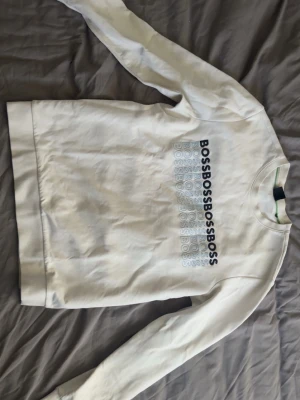 Vit Hugo Boss sweatshirt  - Vit sweatshirt med rund hals och långa ärmar. Framsidan har ett upprepat BOSS-tryck i svart och grått. Tröjan har en klassisk passform och ribbade muddar vid ärmslut och nederkant. Perfekt för en clean och stilren look. Storlek S men passar mig på 186cm och 78kg.