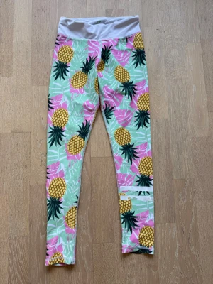 Färgglada leggings/träningsbyxor med ananasmönster - Säljer ett par leggings/träningsbyxor med tropiskt ananasmönster i gult, grönt och rosa. Viss slitning se bild. 