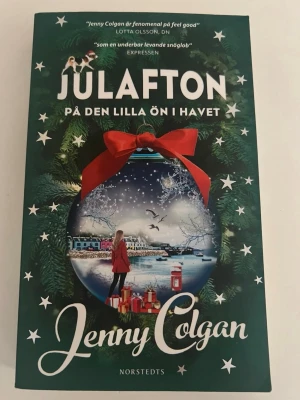 Julafton på den lilla ön i havet - En mysig roman av Jenny Colgan med julig känsla, grön omslag med vintermotiv och en stor julkula med röd rosett. Perfekt för dig som gillar romantik och feelgood i vintrig miljö. Utgiven av Norstedts förlag.