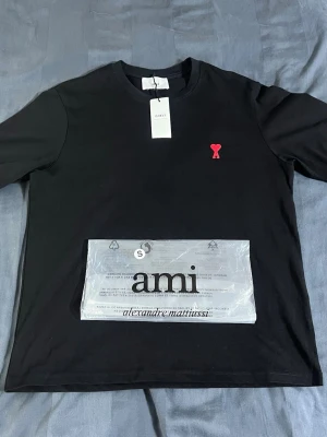 Svart t-shirt från Ami Paris - Svart t-shirt från Ami Paris med klassisk passform och rund hals. På bröstet sitter Ami:s ikoniska röda hjärtemblem. Tillverkad i mjuk bomull som känns skön mot huden. Perfekt till jeans eller shorts för en clean look.OBS stoler s men passar som M