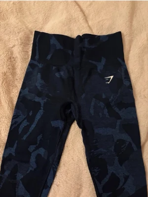 Mörkblå camo leggings från Gymshark -  Snygga mörkblå leggings från Gymshark med camomönster och hög midja. Stretchigt material som sitter skönt och formar sig efter kroppen. Perfekta för träning eller chill. Gymshark-logga på låret för extra stilpoäng .Använda ett fåtal gånger hemma men aldrig på gymmet. I fint skick. Orginalpris runt 400kr 
