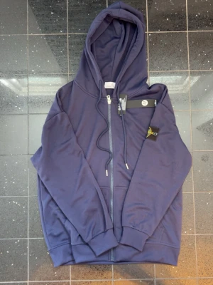 Mörkblå hoodie från Stone Island - Snygg mörkblå hoodie från Stone Island med dragkedja framtill och klassisk logotyp på vänster ärm. Hoodien har justerbar huva med dragsko och ribbade muddar. Perfekt för en avslappnad och trendig stil.