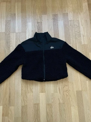 Svart teddyjacka från Alo - Säljer en svart teddyjacka från Alo med hög krage och dragkedja framtill. Jackan har en kontrasterande slät panel upptill och mjukt, fluffigt teddy-material på resten av jackan. Perfekt för att hålla värmen och se stilren ut.