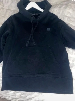 Svart hoodie från Acne Studios - Säljer en svart hoodie från Acne Studios med klassisk huva och dragsko. Hoodien har en stor magficka och en liten Acne Studios-logga på bröstet. Tillverkad i mjukt material som känns skönt mot huden. Perfekt för en avslappnad och stilren look.