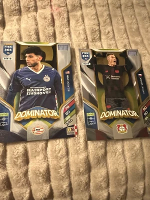 FIFA 365 Dominator fotbollskort - Två FIFA 365 Dominator samlarkort med fotbollsspelare från PSV Eindhoven och Bayer Leverkusen. Kortens design är färgstark med guld, blått och svart, och visar Ricardo Pepi och Patrik Schick i matchtröjor. Perfekt för dig som gillar att samla på fotbollskort.