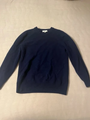 Mörkblå stickad tröja - En enkel och stilren mörkblå stickad tröja med rund halsringning och långa ärmar. Tröjan har ribbade muddar vid ärmslut och nederkant. Perfekt att matcha med jeans eller kjol för en clean look.