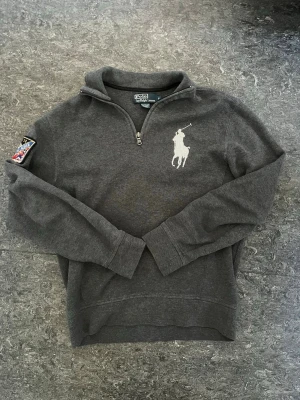 halvzip från Ralph Lauren - snygg och unik mörkgrå sweatshirt med halv dragkedja från Ralph Lauren. Stor vit polospelare broderad på bröstet och en färgglad patch med texten '1933-34 Ralph Lauren' på ärmen. Skön passform och ribbade muddar vid ärmslut och nederkant.