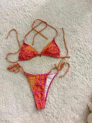 Färgglad bikini med knyt och mönster - Säljer en superfin bikini i starka färger med orange och rosa mönster. Överdelen har trekantskupor och knyt i nacken och ryggen, medan underdelen har höga ben och knyt i sidorna. Perfekt för stranden eller poolen!