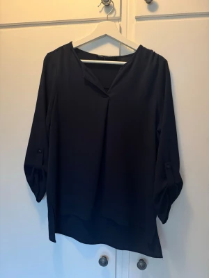 Mörkblå lös blus med knappdetalj - Stilren mörkblå blus i ett mjukt, lätt material. Blusen har V-ringning och trekvartsärmar med knappdetalj som ger en avslappnad look. Passar perfekt till jeans eller kjol för en enkel men snygg outfit. Finns dock små fläckar baktill, kanske går att få bort