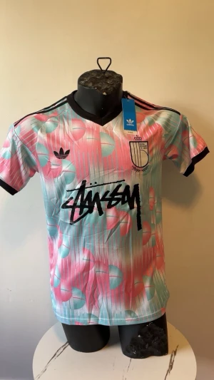 Adidas x Stüssy färgglad sporttröja - Säljer en unik sporttröja från Adidas i samarbete med Stüssy. Tröjan har ett färgstarkt mönster i rosa, turkos och vitt med svarta detaljer, V-ringning och korta ärmar. Adidas-logga och Stüssy-tryck framtill. Perfekt för dig som gillar streetwear och sportig stil.