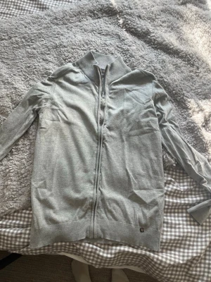 Ljusgrå ziptröja från INDICODE - Snygg ljusgrå ziptröja från INDICODE med hög krage och ribbade muddar. Tröjan har hel dragkedja framtill och är tillverkad i mjukt material som passar perfekt till lager på lager. Enkel och stilren design utan tryck.