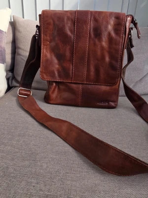 Brun axelväska i äkta läder - Snygg brun axelväska i äkta läder med justerbar axelrem och lock. Väskan har en stilren design med vertikala sömmar och en praktisk dragkedjeficka på baksidan. Mått 24x29x6