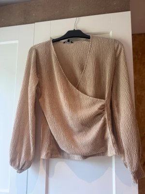 Beige omlottopp med lång ärm - Säljer en beige omlottopp med lång ärm och lätt glansigt, strukturerat tyg. Toppen har v-ringning och en avslappnad passform som ger en snygg siluett. Perfekt att styla med jeans eller kjol för en trendig look. Storlek 36/38