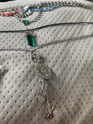 Mixade halsband med färgglada detaljer - Säljer ett set med tre halsband: ett med stora färgglada stenar i blått, grönt och rött samt en stor klar kristall, ett med silverkedja och grön smaragdliknande sten, och ett med silverfärgade mynt och kedjor. Perfekt för dig som gillar statement-smycken och att mixa stilar.