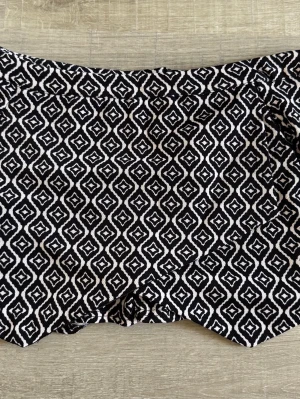 Svarta shorts med vit geometrisk mönster - Säljer ett par svarta shorts med vitt geometriskt mönster. Shortsen har en rak passform och är fodrade med ett slätt svart tyg på insidan. Perfekta för dig som vill sticka ut lite extra i sommar!