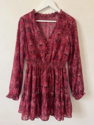 Röd blommig kortklänning med volang - Supersöt röd kortklänning med blommigt mönster, v-ringad hals och långa ärmar. Klänningen har smock i midjan och volangkant nedtill för en flowy look. Perfekt för vår och sommar, till sneakers eller boots.