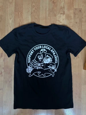 Malmö FF BU Svart t-shirt AppaJävel - Cool svart t-shirt med vitt tryck framtill som föreställer en apa och en polis med texten 'Support Your Local Apajävel'. Klassisk rund hals och korta ärmar. Perfekt för dig som vill sticka ut med en unik och rolig design.