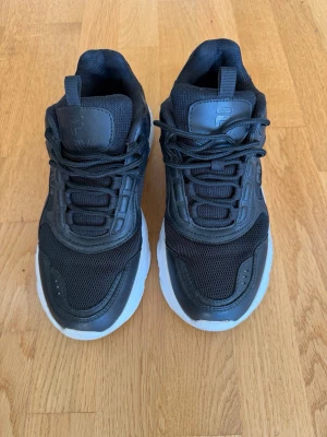 Sneakers från Fila - Svarta sneakers från Fila med chunky vit sula och ovandel i mesh och syntetmaterial. Skorna har snörning och en sportig vibe, perfekt för dig som gillar streetstyle. Fila-logga syns på sidan och plösen.
