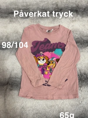 Rosa långärmad Paw Patrol t-shirt - Söt rosa långärmad t-shirt med Paw Patrol-tryck framtill. Tröjan har rund halsringning och motiv i lila, rosa och turkos. Perfekt för alla små fans av Paw Patrol.🌸påverkat tryck. Storlek 98/104