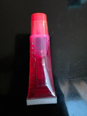 Rosa Magic Gloss läppglans - Säljer ett läppglans i tub med transparent rosa färg och texten 'MAGIC GLOSS' på framsidan. Tuben har en praktisk skruvkork i matchande rosa och är gjord av mjuk plast som är enkel att klämma ut produkten ur. Perfekt för dig som vill ha glansiga läppar med en hint av färg.