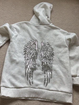 Ljusgrå hoodie med stora vingar där bak - Säljer en ljusgrå hoodie med huva På ryggen finns ett stort motiv av änglavingar i silvriga paljetter som verkligen sticker ut. Hoodien har en klassisk känguruficka framtill och ribbade muddar vid ärmar och nederkant. Perfekt för dig som gillar statement-plagg.