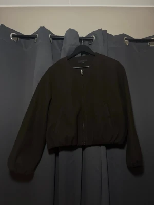 Mörkbrun bomberjacka från ZARA - Snygg mörkbrun bomberjacka från ZARA med dragkedja framtill och två fickor. Jackan har en enkel, stilren design med rundad krage och resår i midjan. Tillverkad i ett mjukt tyg som ger en avslappnad look. Använd 1 gång, inget slitage.