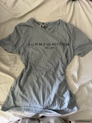 Ljusblå Tommy Hilfiger t-shirt - Säljer en ljusblå t-shirt från Tommy Hilfiger med tryckt logga och texten 'EST. 1985' på bröstet. Klassisk rund halsringning och korta ärmar. Perfekt för en avslappnad och stilren look, har dock en fläck på baksidan vid nacken