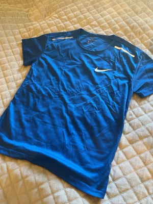 Blå Nike t-shirt med vit logga - Snygg blå t-shirt från Nike med vit swoosh-logga på bröstet och reflekterande detaljer vid axlarna. Tillverkad i ett lätt och skönt material som passar perfekt till träning eller vardagsstil. Klassisk rund hals och korta ärmar.