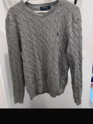 Grå kabelstickad tröja Polo Ralph Lauren - Säljer en grå kabelstickad tröja från Polo Ralph Lauren med rund hals och ribbade muddar. tillverkad i mjukt material som passar perfekt för kyliga dagar.