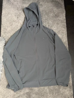 Nike unlimated vindjacka  - Säljer en stilren grå hoodie från Nike med hel dragkedja framtill och huva. Hoodien har långa ärmar, diskret Nike-logga på bröstet och elastiska muddar vid ärmsluten. Perfekt för en chill och sportig look.