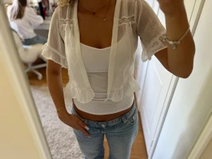 Vit transparent blus med volang - Söt vit blus i transparent material med volangdetaljer längs ärmslut och nederkant. Blusen har korta ärmar och fina spetsinslag upptill. Perfekt att styla över ett linne eller topp för en trendig lager-på-lager-look.