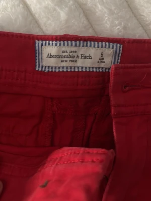 Röda shorts från Abercrombie & Fitch - Snygga röda shorts från Abercrombie & Fitch med klassisk passform och uppvikta benslut. De har dragkedja och knapp framtill samt bälteshällor. Perfekta för varma dagar och enkla att matcha med olika toppar.