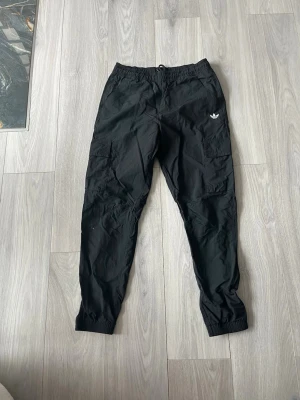 Svarta cargobyxor från Adidas - Snygga svarta cargobyxor från Adidas med elastisk midja och muddar vid benslut. Byxorna har flera praktiska fickor på sidorna och en vit Adidas-logga på vänster sida. Tillverkade i ett lätt och slitstarkt material som ger en sportig look.