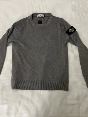 Grå stickad tröja från Stone Island - Grå stickad tröja från Stone Island med klassisk rund halsringning och ribbade muddar. Tröjan har den ikoniska Stone Island-loggan på vänster ärm. Längd från axelhöjd till midjan: 58-60 cm. Pris kan diskuteras😊
