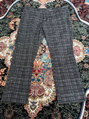 Grå rutiga kostymbyxor bootcut - Snygga grå kostymbyxor med vitt rutmönster och bootcut-ben. Byxorna har klassisk passform, hällor för skärp och stängs med knapp och dragkedja. Materialet ser ut att vara en blandning av polyester och viskos för en mjuk känsla.