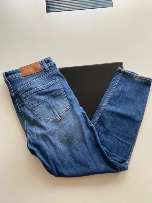 Blå raka jeans i klassisk modell - Säljer ett par blå jeans i klassisk rak modell med fem fickor och bälteshällor. Jeansen har en snygg tvättad look och är tillverkade i denim. Passar perfekt till en avslappnad stil och är ett måste i garderoben.
