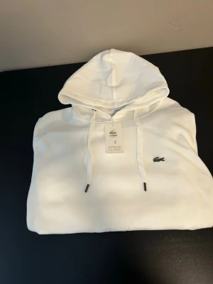 Vit hoodie från Lacoste med huva - Säljer en clean vit hoodie från Lacoste med klassisk huva och dragsnören. Ikoniska krokodilloggan broderad på bröstet. Hoodien har en relaxed fit och är gjord i mjukt material som känns skönt mot huden. Perfekt för dig som gillar stilren och enkel design.