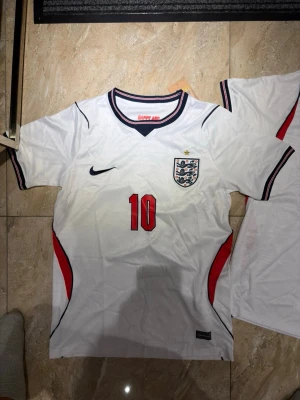 England fotbollströja Bellingham 10 - Säljer en vit England fotbollströja från Nike med nummer 10 och Bellingham på ryggen. Tröjan har röda och svarta detaljer, broderad landslagssköld och Nike-logga på bröstet. Kortärmad modell med sportig passform, perfekt för match eller träning.