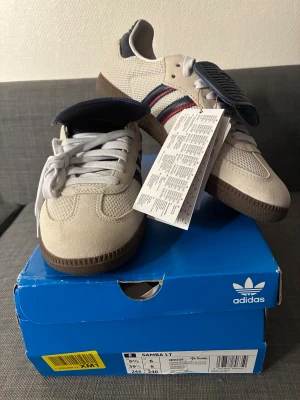 Adidas Samba beige sneakers NYA - Helt nya Adidas Samba LT sneakers i beige med klassiska mörkblå och röda ränder på sidan. Exklusiv modell som är slutsåld i de flesta storlekar. Skorna har vit snörning, brun platt sula och en mix av mocka och mesh-material på ovandelen. Ikonisk retrostil med rund tå och detaljer i guld vid loggan. 