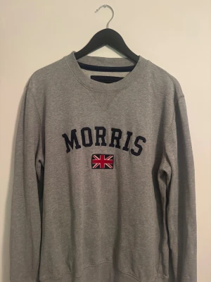 Grå sweatshirt från Morris - Klassisk grå sweatshirt från Morris med mörkblå text och en broderad brittisk flagga på bröstet. Tröjan har rund halsringning och ribbade muddar vid ärmslut och nederkant. Perfekt för en avslappnad och stilren look.