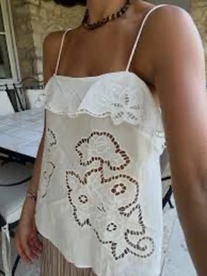 Vit spetslinnetopp med volang - Söt vit linnetopp med tunna axelband och volangkant upptill. Toppen har broderade spetsdetaljer och blommönster som ger en romantisk vibe. Perfekt för varma dagar och sommarens alla äventyr.