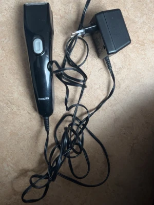 Svart hårtrimmer från Philips - En svart hårtrimmer från Philips med sladd och tillhörande laddare. Trimmern har en enkel design med en vit knapp på framsidan och metallblad upptill. Perfekt för att fixa frisyren hemma.
