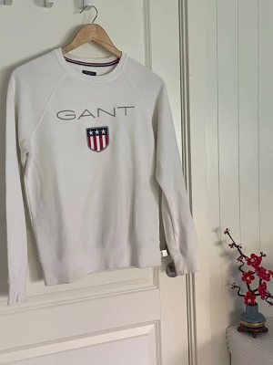Vit sweatshirt från GANT XS - Säljer en vit sweatshirt från GANT med broderad logga och amerikanskt inspirerat sköldmärke på bröstet🤩Tröjan har rund halsringning och ribbade muddar vid ärmslut och nederkant✌️