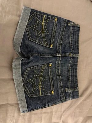 Mörkblå jeansshorts med stjärndetaljer - Snygga mörkblå jeansshorts med uppvikta benslut och coola stjärndetaljer i gult på bakfickorna. Shortsen har klassisk femficksdesign, kontrastsömmar och guldfärgade nitar. Perfekta för sommardagar och enkel att matcha med olika toppar.
