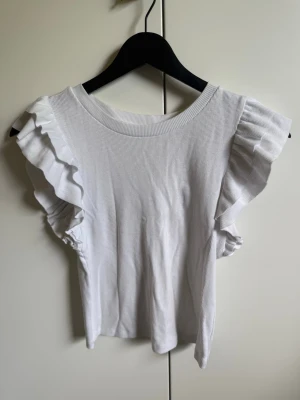 Vit ribbad topp med volangärmar - Snygg vit ribbad topp med volangdetaljer på ärmarna. Toppen har rund halsringning och är ärmlös med breda volangprydda axlar som ger en trendig look. Perfekt att styla till jeans eller kjol för en fräsch outfit.