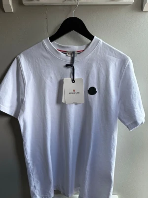 Vit Moncler t-shirt med logga - Snygg vit t-shirt från Moncler med svart logomärke på bröstet. Klassisk rund halsringning och korta ärmar. Tillverkad i mjuk bomull för en skön känsla. Perfekt till jeans eller shorts för en clean look.