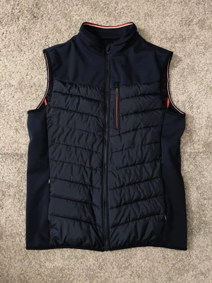 Svart dunväst med röda detaljer - Snygg svart dunväst med quiltad framsida och röda detaljer längs dragkedja och insida. Västen har hög krage, bröstficka med dragkedja och två innerfickor. Materialet är glansigt och lätt, perfekt för lager-på-lager. Passar dig som vill ha en stilren och sportig look.